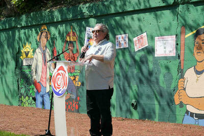 A Hispanic male stands at a podium with a large green mural behind him, depicting baseball players.