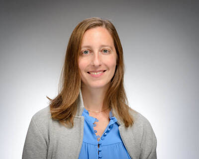 A smiling woman with light brown hair and blue eyes. She wears a blue button-front blouse and a gray cardigan, standing against a plain gray background.